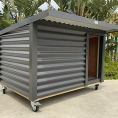 a small metal structure with wheels and a door on the side, in front of some palm trees