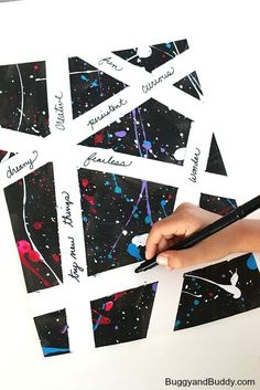 a child's hand holding a pencil and writing on some black paper with colorful paint splattered all over it