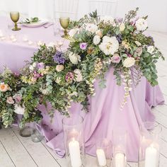 an arrangement of flowers and greenery on a table with candles in front of it