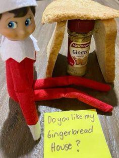 an elf is sitting in front of a gingerbread house with a sign that says do you like my gingerbread house?