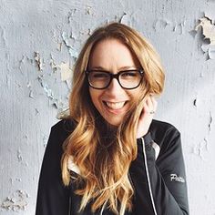 a woman wearing glasses is smiling and talking on her cell phone while standing in front of a wall with peeling paint