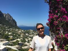 a woman standing on top of a hill next to purple flowers and the ocean in the background