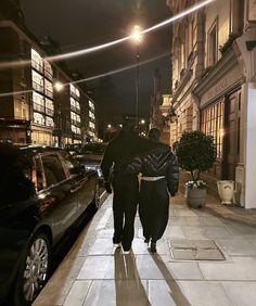 two people walking down the street at night with cars parked on the side walk and buildings in the background