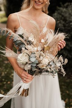 a woman in a white dress holding a bouquet with dried flowers and feathers on it