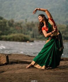 a woman in a green and orange sari dancing on the shore of a river