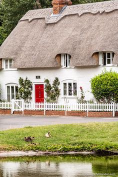 a white house with a red front door and thatched roof next to a body of water