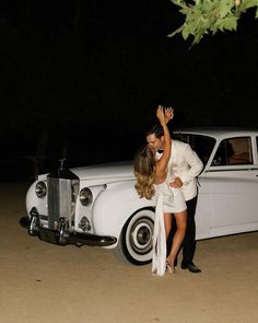 a bride and groom kissing in front of an old white car at night with their arms around each other