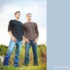 two men standing next to each other in front of tall grass and blue sky with clouds