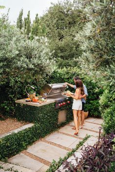 a woman standing next to an outdoor grill in the middle of some bushes and trees