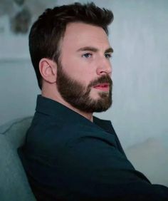 a man with a beard sitting on a couch looking off to the side and wearing a black shirt