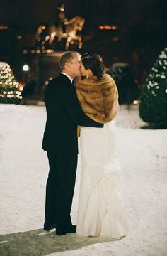 a bride and groom kissing in the snow at their winter wedding reception with lights on behind them