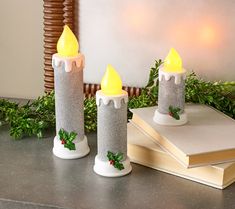 two christmas candles with holly decorations on them sitting next to a stack of books and greenery