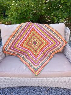 a white wicker chair with a colorful pillow on it's back sitting in front of a bush