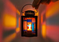 a hand holding a small lantern with a lit candle in it's center against a multicolored background