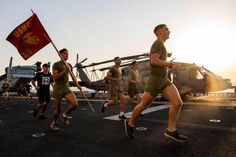 men running on an aircraft carrier with a flag in the foreground and a helicopter behind them