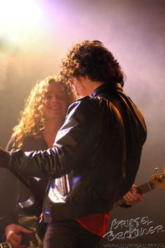 two people standing next to each other with guitars in their hands and one person holding an electric guitar