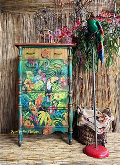 a colorful painted dresser next to a basket with flowers on it and a parrot sitting on top