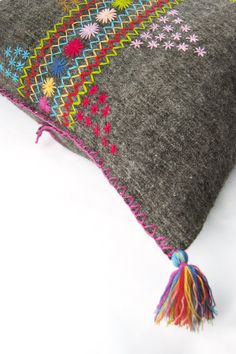 a gray pillow with multicolored flowers and tassels on the front, sitting on a white surface