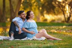 a man and woman are sitting on the grass in front of trees with their arms around each other