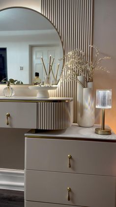 a white dresser topped with a mirror and vase filled with flowers next to a lamp