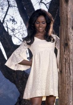 a woman standing next to a tree wearing a white dress with bell sleeves and high heels