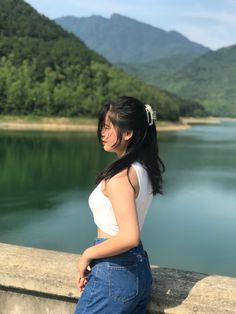 a woman standing on the edge of a wall next to a body of water with mountains in the background