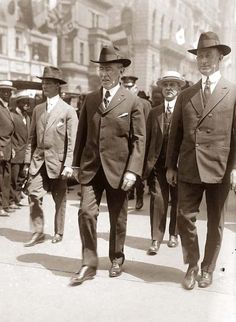 several men in suits and hats walking down the street with other people behind them wearing hats