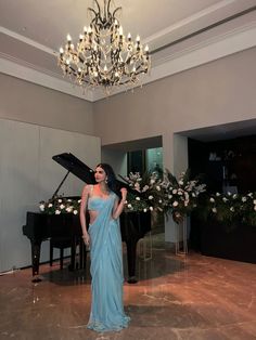 a woman in a blue dress standing next to a grand piano and chandelier