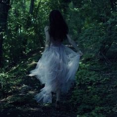 a woman in a white dress walking through the woods with her back to the camera