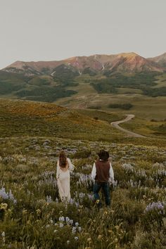 two people are walking through a field with wildflowers and mountains in the background