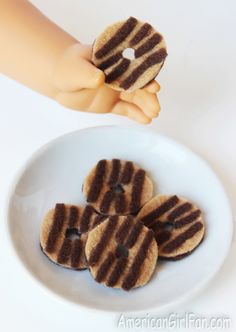 a white plate topped with chocolate covered donuts next to a doll's hand