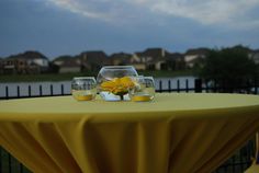 a yellow table cloth with two glasses and a flower on it, in front of a lake