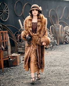 a woman in a fur coat and hat walks down a dirt road near an old building