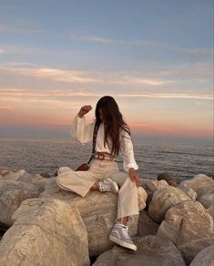 a woman sitting on rocks near the ocean at sunset with her hair blowing in the wind