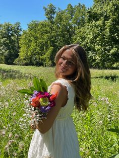 a girl in a white dress holding flowers and smiling at the camera while standing in a field