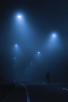 a man walking down a street at night in the fog with three lights on either side of him