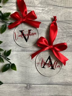 two personalized glass ornaments with red ribbon and bow on them, sitting next to greenery