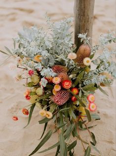 an arrangement of flowers and greenery on the sand at the beach in front of a wooden pole