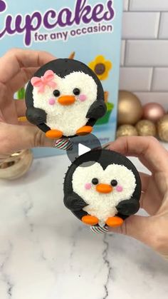 two small penguins are being held up by someone's hands in front of a box of cupcakes