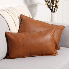 two brown leather pillows sitting on top of a gray couch next to a vase with flowers