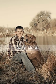 a man sitting next to a brown dog on top of a grass covered field in front of a lake