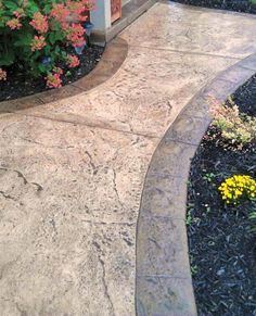 an outdoor walkway in front of a house with flowers on the side and bushes around it