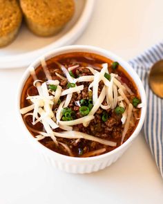 a bowl of chili and cheese next to some cookies on a white tablecloth with two spoons