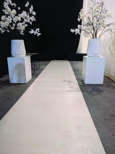 a white carpeted aisle with vases and flowers on each side in front of a black backdrop