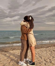 two people standing on the beach hugging each other and looking out at the ocean under a cloudy sky