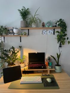 a desk with a laptop, keyboard and plants on it in front of a clock