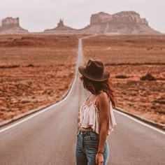 a woman standing in the middle of an empty road wearing jeans and a hat with her hands behind her back