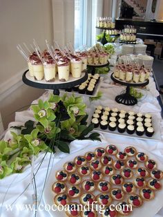 an assortment of desserts are displayed on a long table with white cloth and green flowers