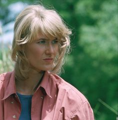 a woman with blonde hair wearing a pink shirt and looking off to the side in front of trees