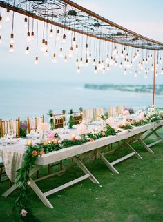 a long table with flowers and lights hanging from it's ceiling in the grass